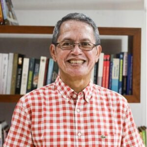 Pak Agus tersenyum di depan rak buku, mengenakan kemeja kotak merah putih sebagai penulis artikel opini tentang banjir dan longsor Sumatra.